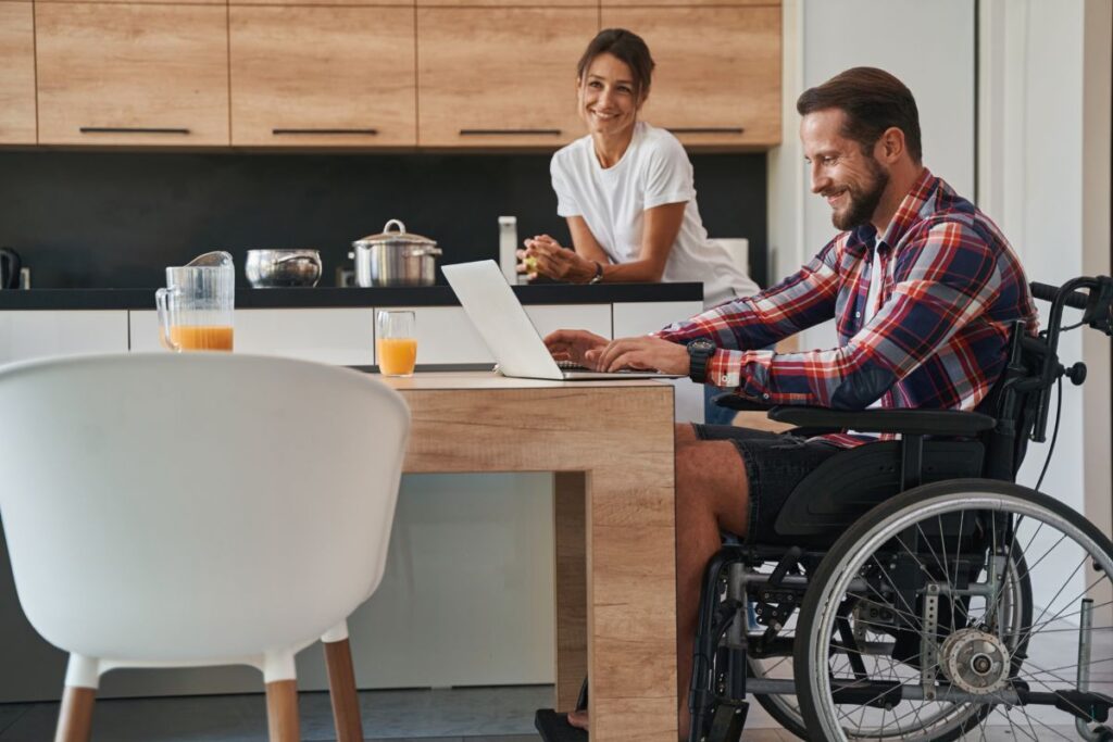 A man in a wheelchair sits at a kitchen table working on a laptop, while his partner is seen in the background.