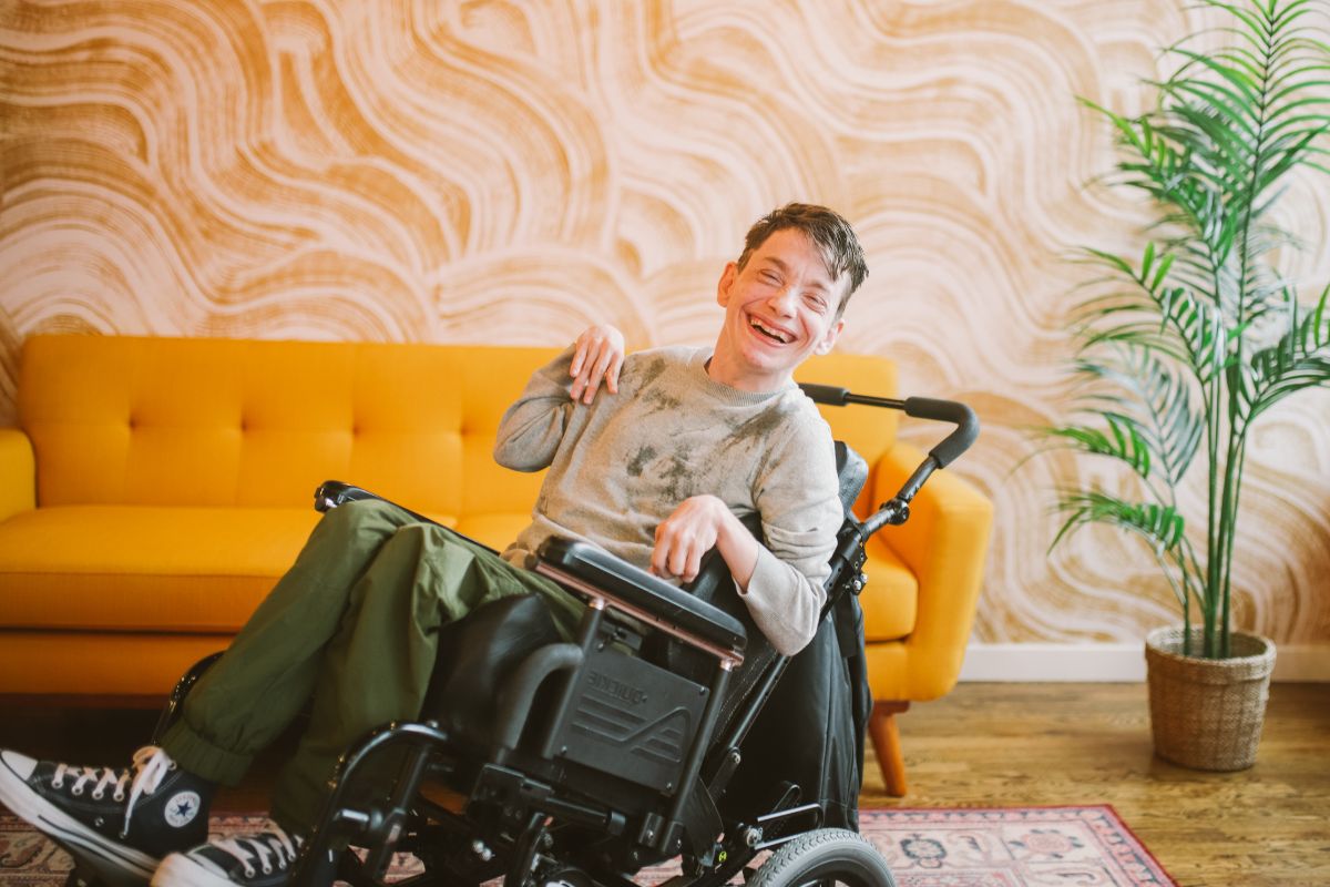 A young man with a disability sits in his wheelchair in front of a yellow couch and living room.