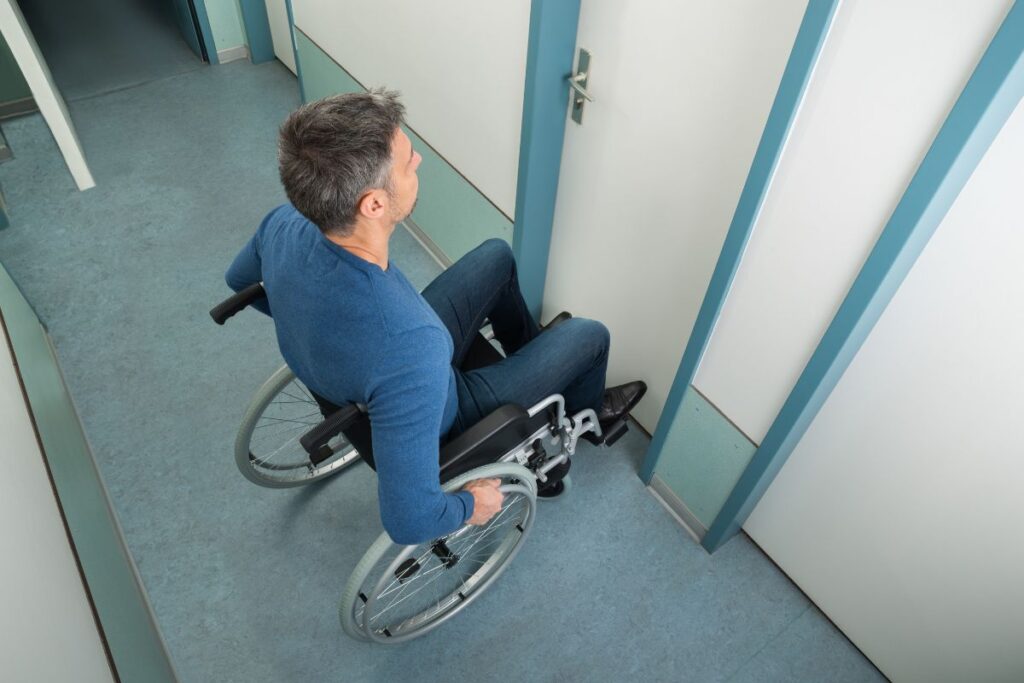 A Caucasian man in a wheelchair is outside a door in a hallway.